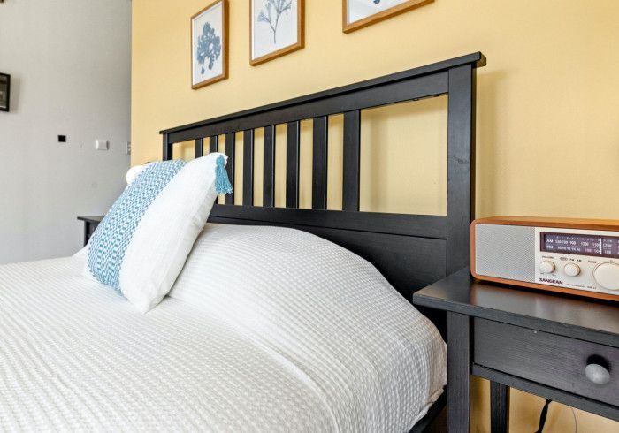 Bedroom with black headboard, white bedding, blue and white pillow, nightstand with radio, and yellow wall.