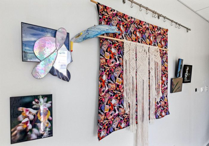 Artwork on a white wall, includes floral photography, a butterfly-shaped stained-glass piece, and a patterned textile with macramé.