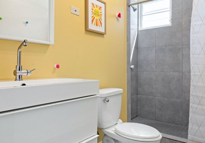 Modern yellow-walled bathroom with white sink, toilet, and grey-tiled shower. Artwork on wall.