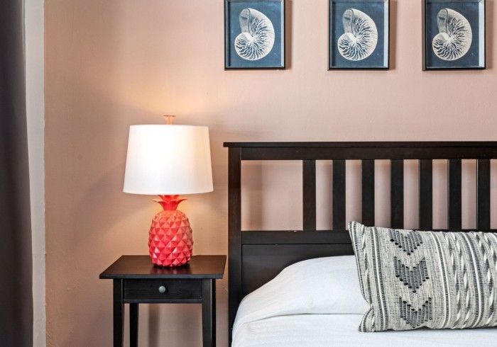 Bedroom with pink wall, dark wood bed, black nightstand with pineapple lamp. Three seashell prints above.