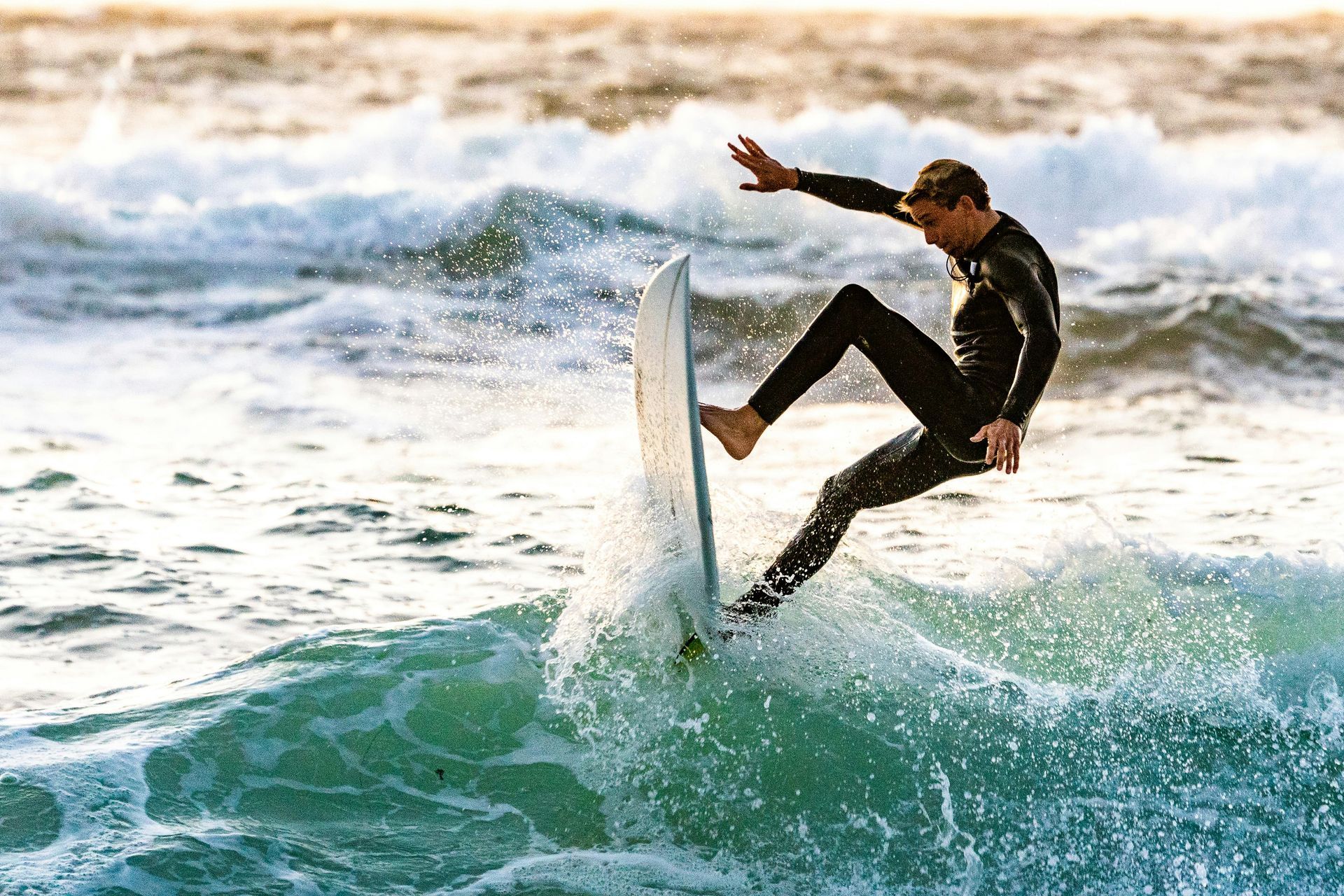 Surfer in black wetsuit carves a wave on a turquoise sea, arm outstretched, at sunset.