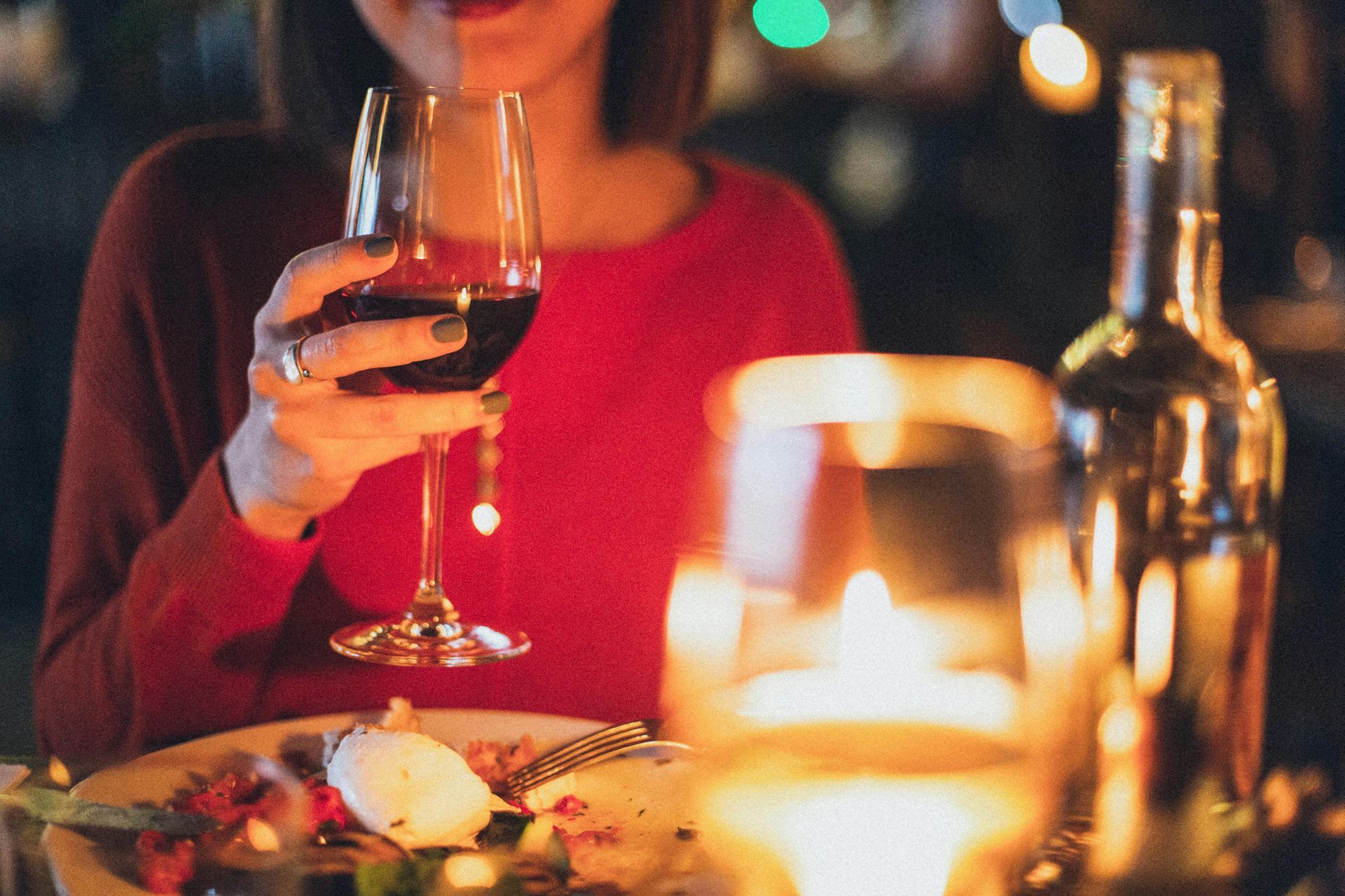 Woman in red sweater holds wine glass at a candlelit dinner. A bottle and another glass are visible.