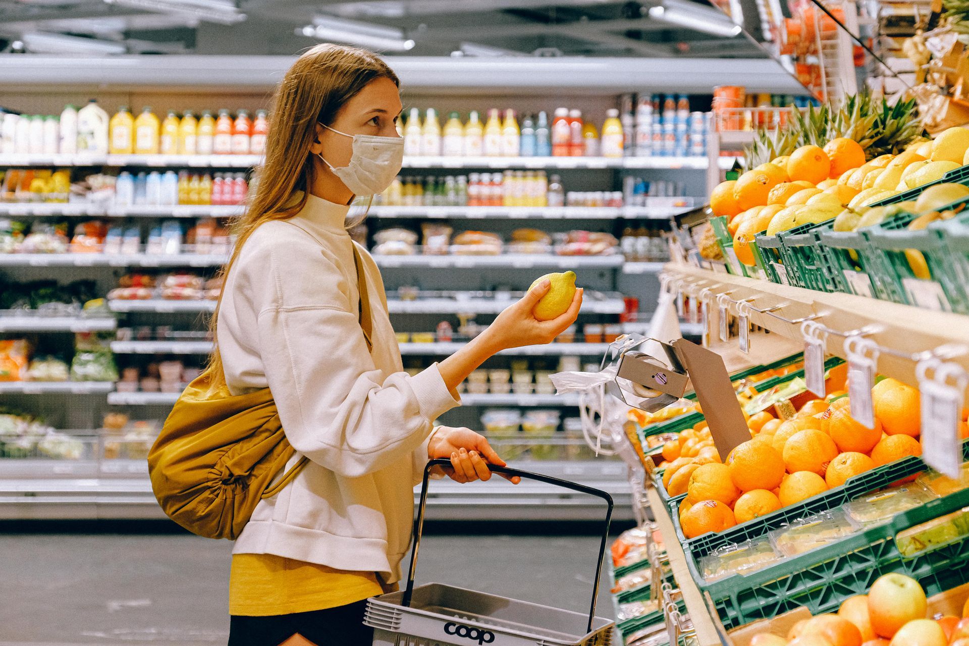 Woman in a face mask shopping for produce in a brightly lit grocery store.