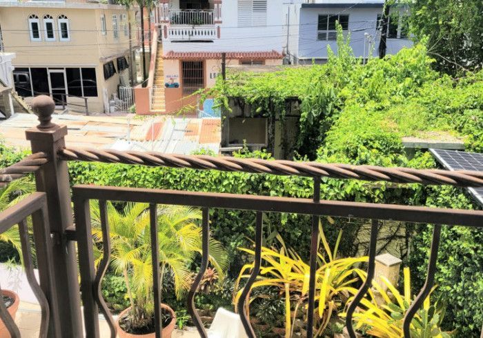 Balcony overlooking street with houses and greenery. Brown railing, plants, sunny.