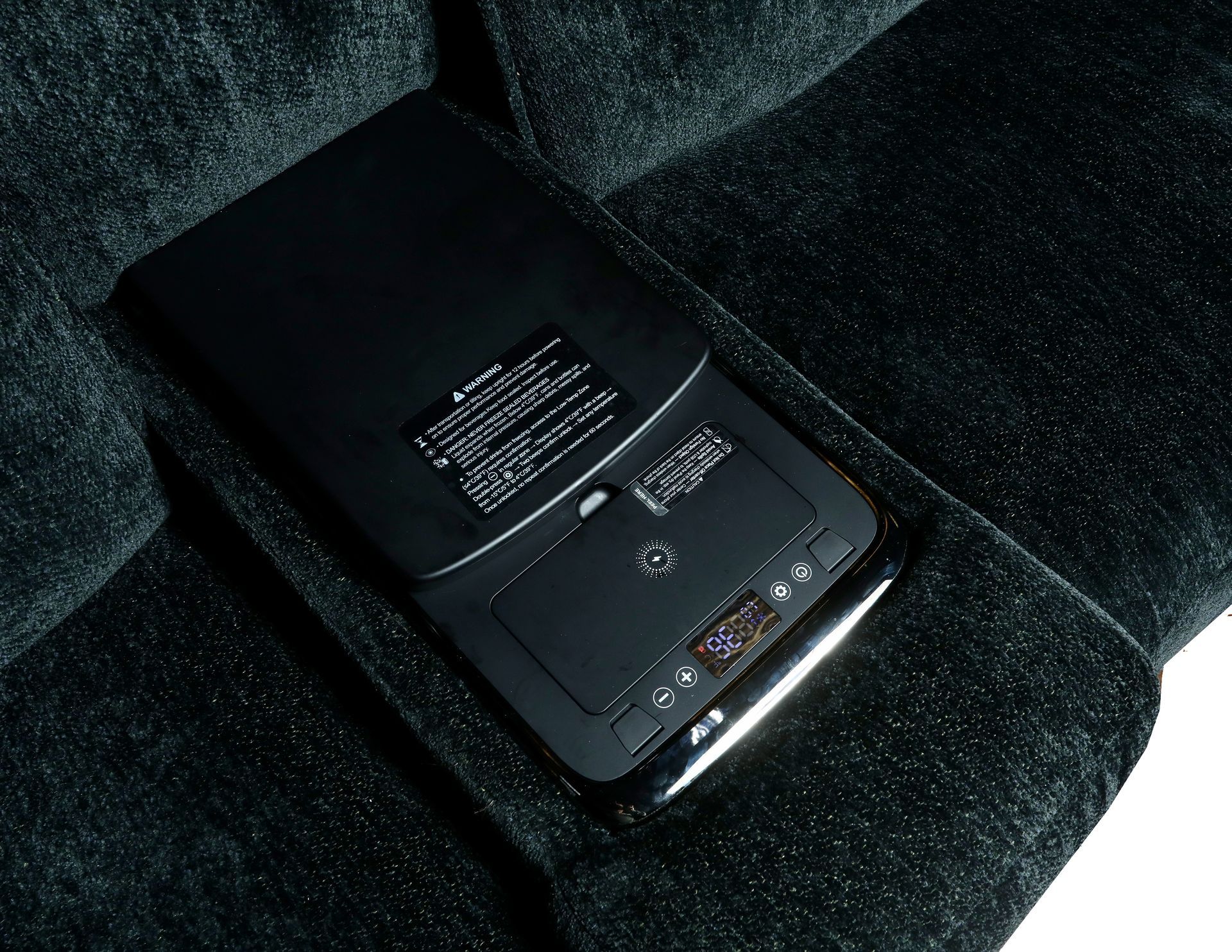 A black, rectangular electronic control panel for a massage chair sits on a dark, textured fabric sofa seat.