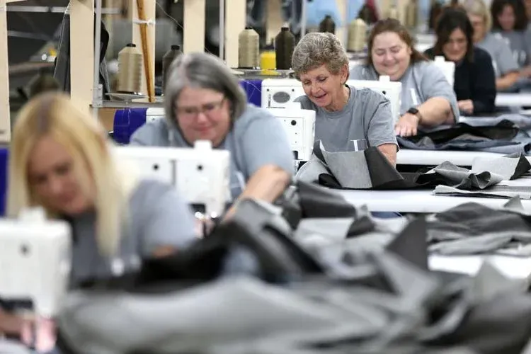 A row of people wearing gray shirts sewing black fabric on industrial machines in a brightly lit factory.