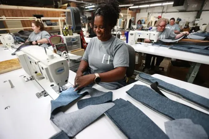 Several people work at industrial sewing machines in a bright factory, focused on stitching pieces of denim fabric.