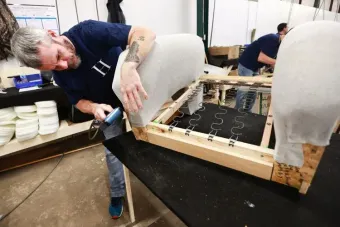 A person in a dark blue shirt uses a staple gun to attach fabric to the wooden frame of a chair in a workshop.