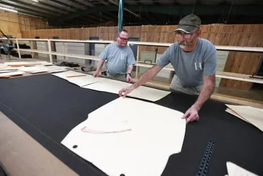 Two workers arrange paper patterns on a long black fabric cutting table in a workshop.
