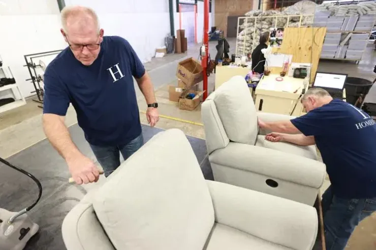 Two employees in matching blue shirts inspect and assemble off-white upholstered recliners in a warehouse setting.
