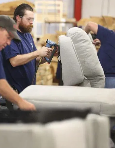 Two workers in an upholstery shop use staple guns to attach light gray fabric to furniture frames.