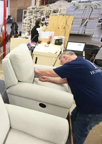 A person in a blue shirt works on the upholstery of a light-colored armchair in a warehouse setting.
