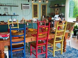 Colorful wooden chairs around a rustic dining table in a bright room with blue rug and garden view