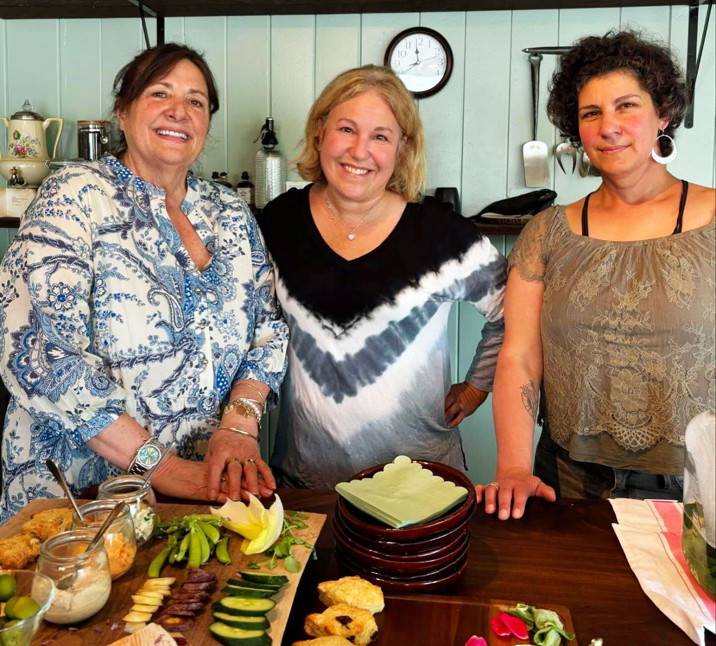 Three women smiling behind a table of food in a cozy kitchen