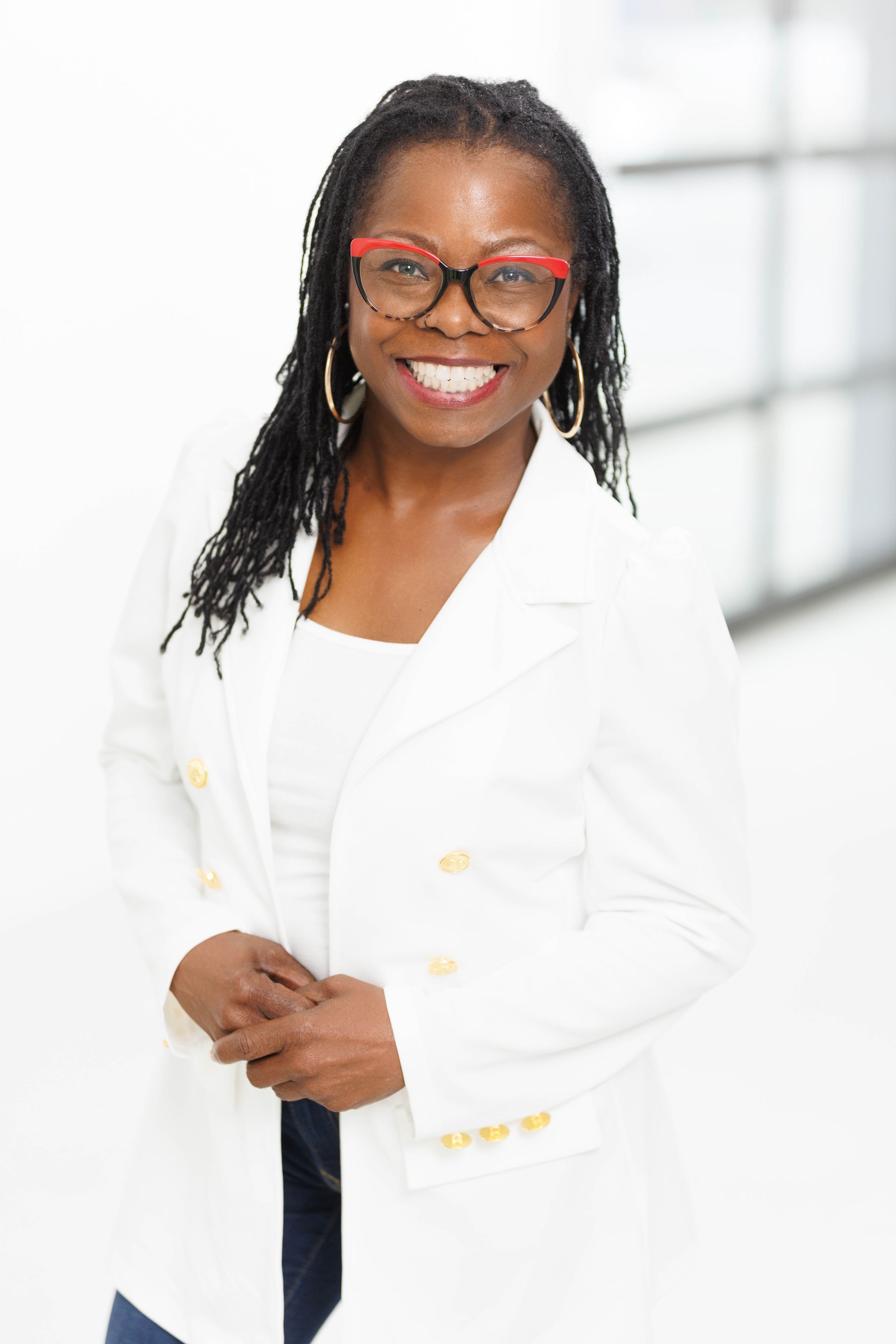 Woman in white blazer, red-rimmed glasses, and smile. Hands clasped, standing indoors near windows.