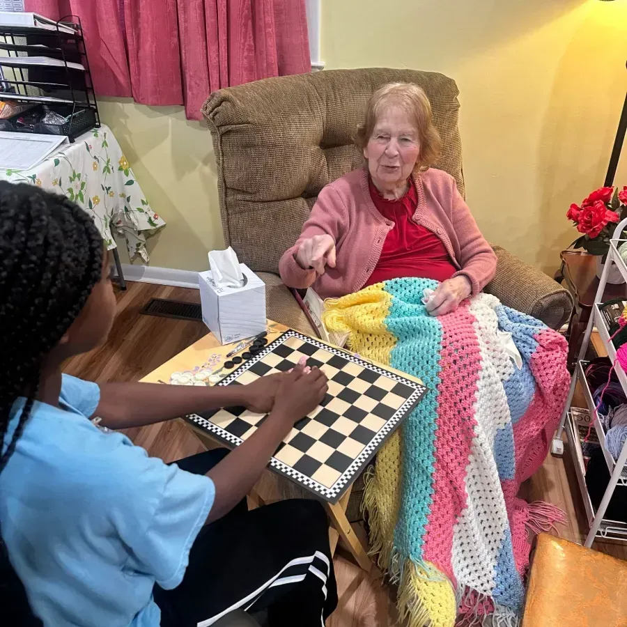 Two people play checkers beside an elderly woman reclining under a colorful crocheted blanket.