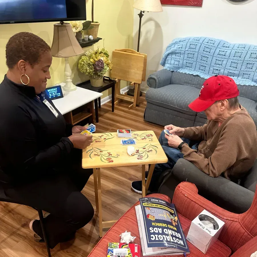 Two people playing a tabletop game in a living room, with cards on a small table and a red chair nearby.