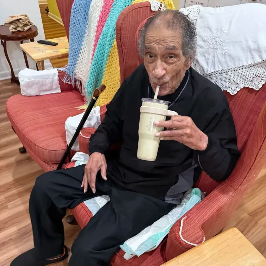 Man sipping a drink with a straw while seated on a red couch with a colorful blanket behind him