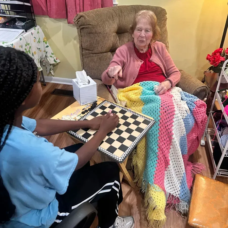 Two people play checkers beside an elderly woman wrapped in a colorful blanket on a chair