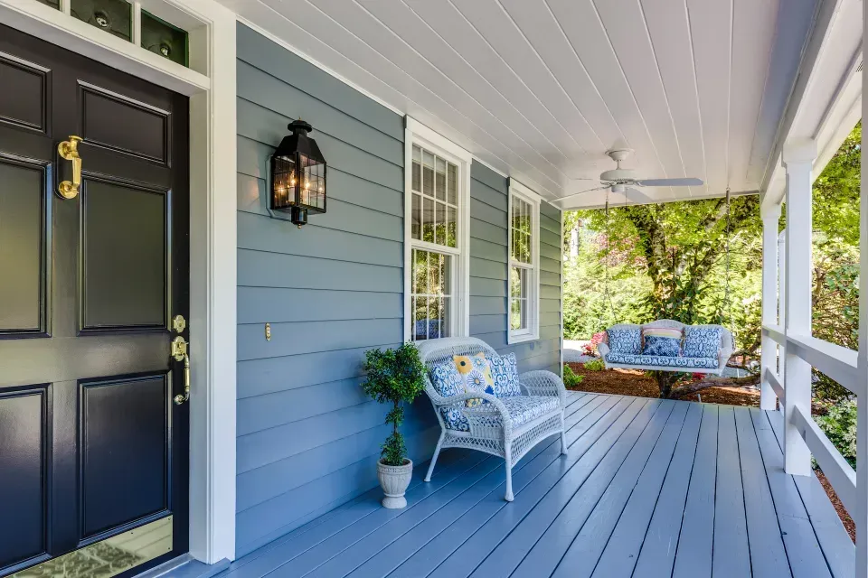 A porch with blue floor and siding with a wicker couch and a black door