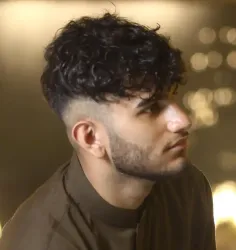 A man with curly hair and a beard is wearing a brown shirt