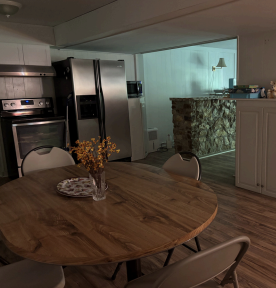 Dining area with round wooden table, chairs, and a kitchen with stainless steel appliances in the background.