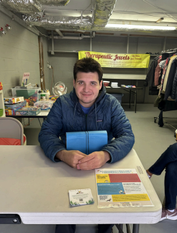 Man sitting at a table indoors, holding a blue folder, with a banner and clothing racks in the background.