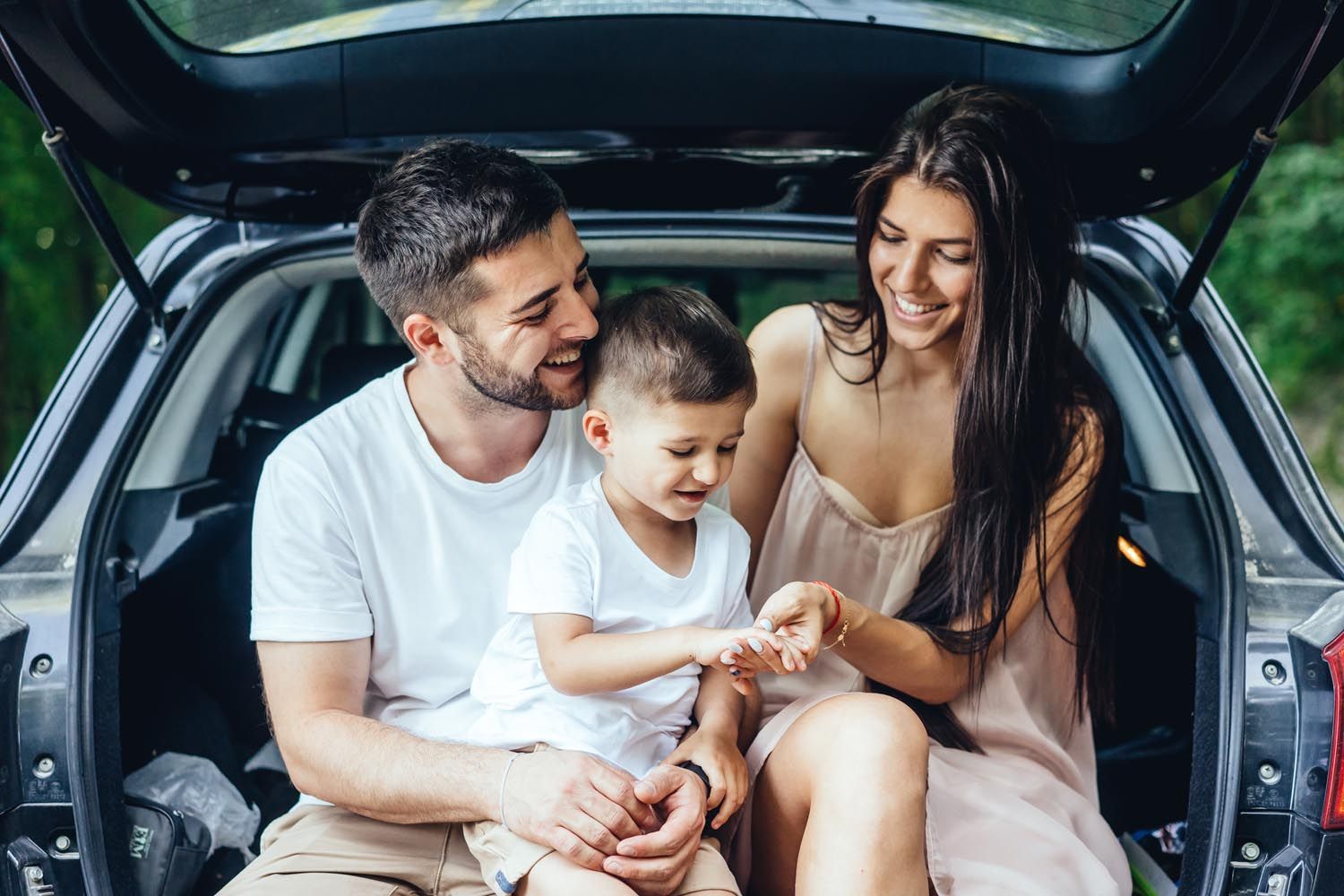 Family of three smiling while sitting in an open car trunk outdoors.