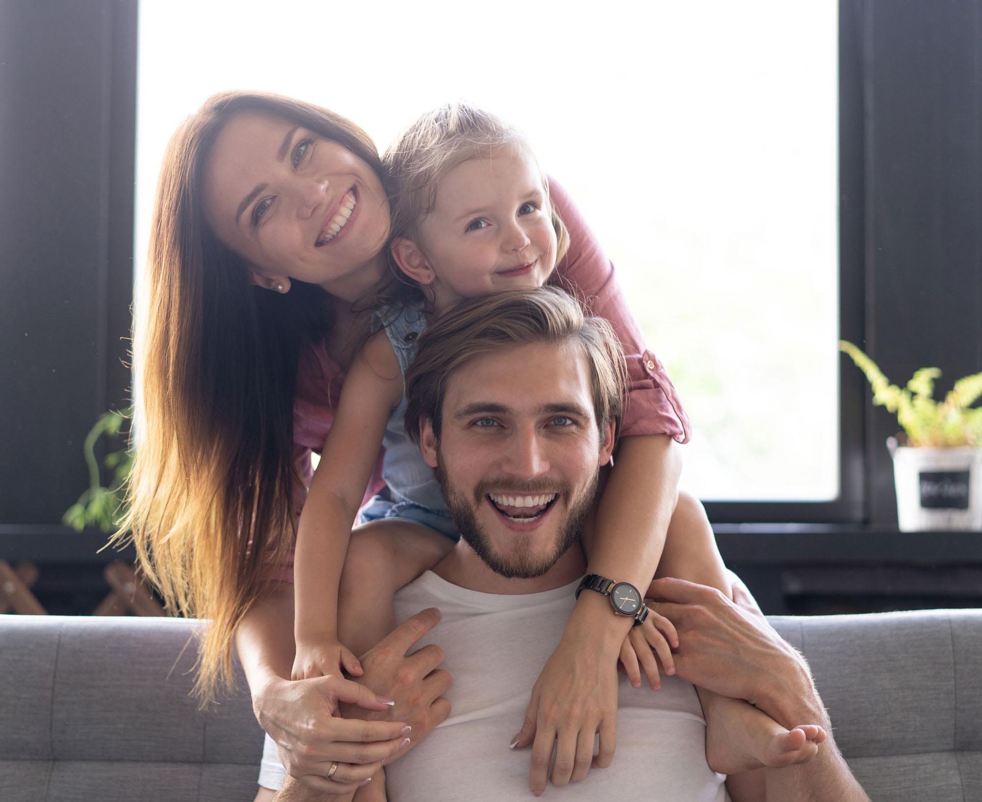 Smiling family of three in a home setting. A woman and child are on the man's shoulders. All are happy.