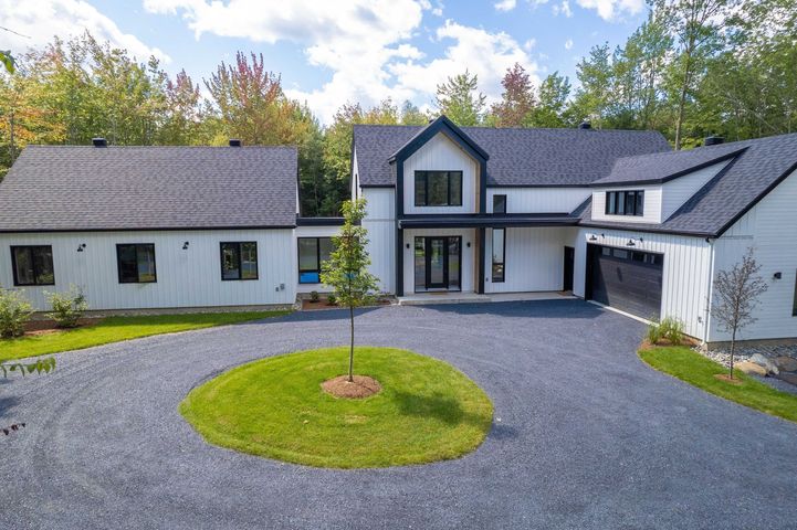 Une maison de ferme moderne blanche à deux étages avec un toit en métal foncé, située derrière une allée circulaire en gravier avec une pelouse centrale.
