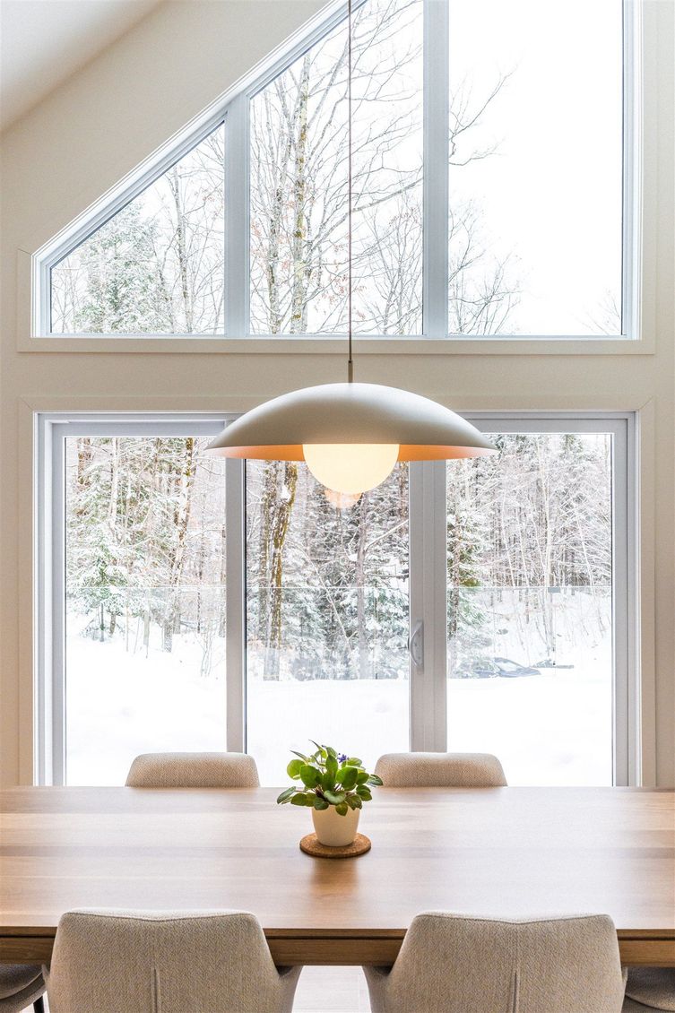 Une table à manger avec une plante en pot, sous une grande suspension lumineuse, devant des baies vitrées donnant sur une forêt enneigée.