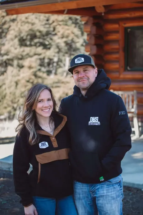 Two people standing outside a log cabin, wearing black hoodies, smiling at the camera.