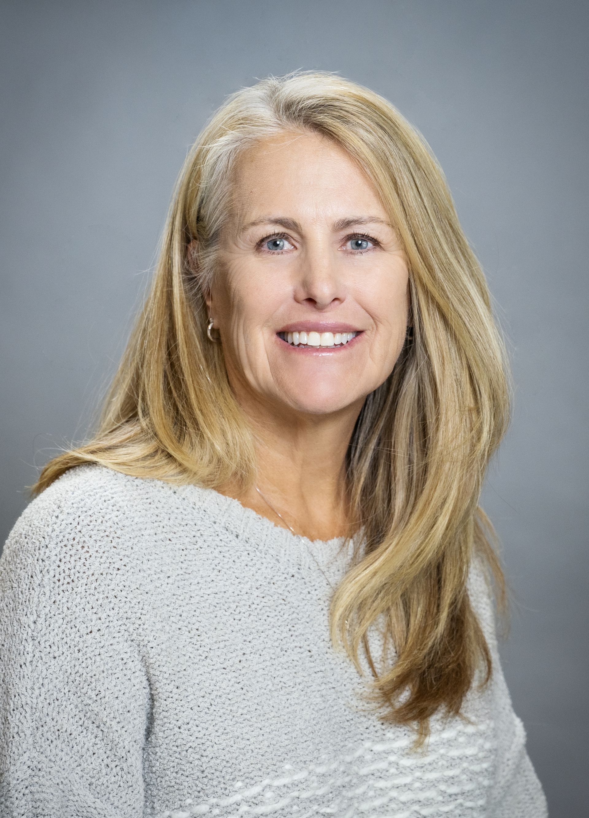 A woman in a pink and brown shirt is smiling in front of trees.