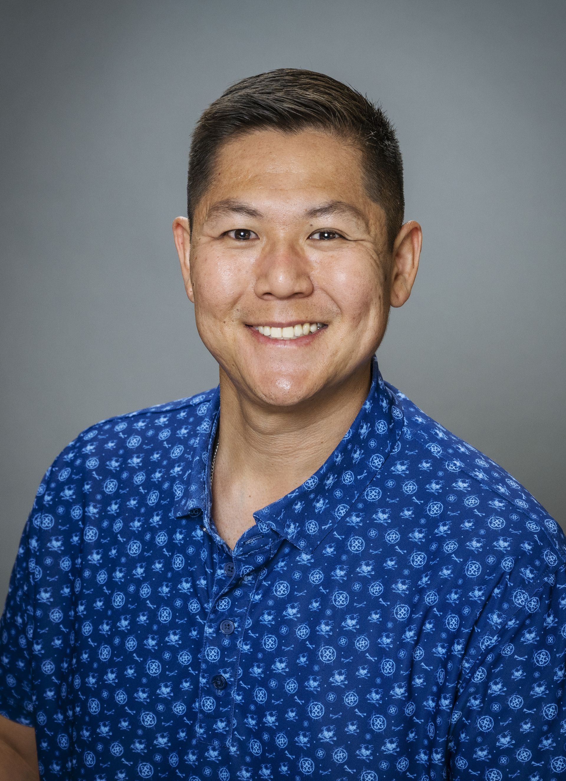 A man in a blue shirt is smiling in front of a forest.