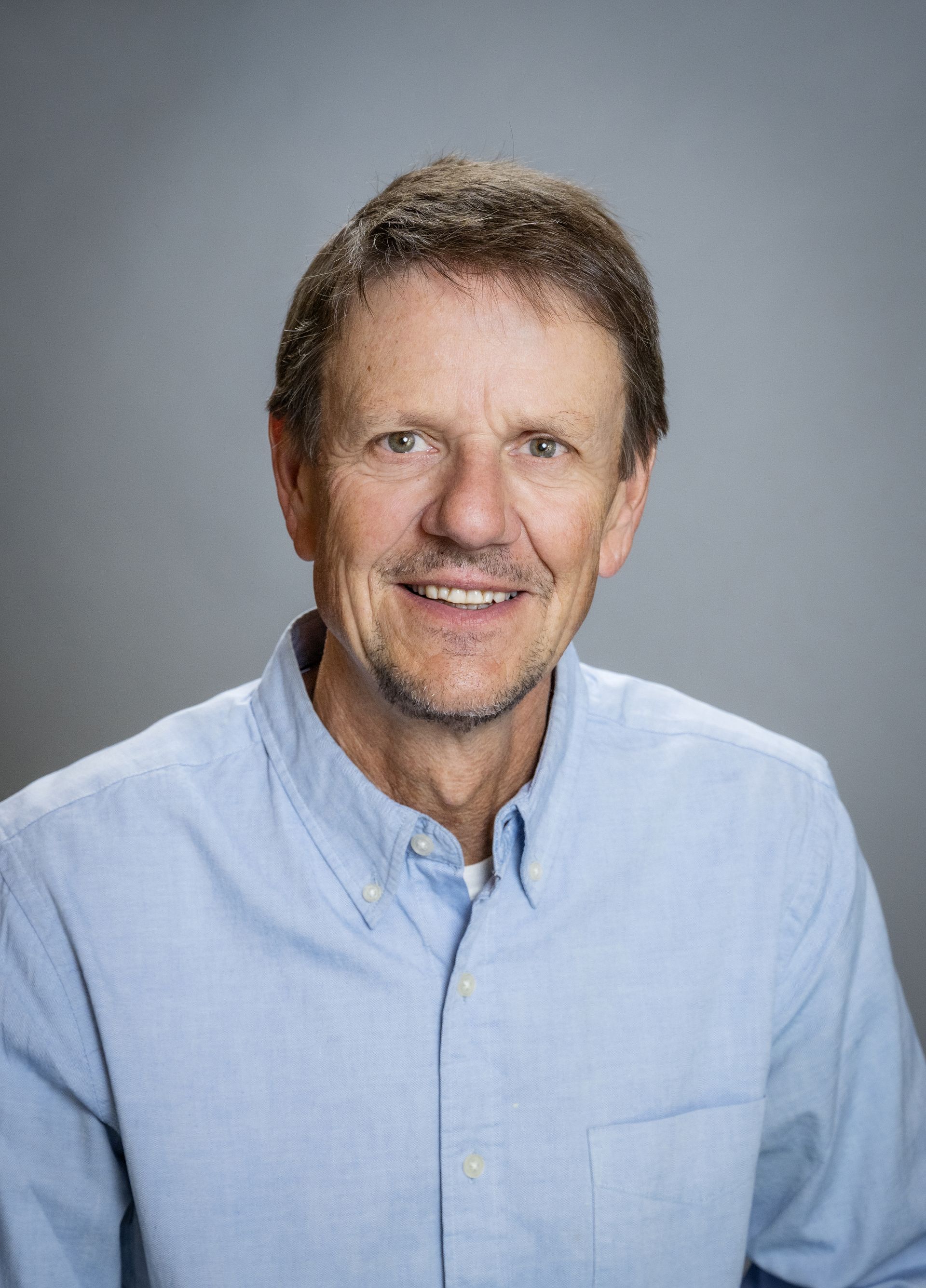 A man in a blue shirt is smiling in front of a bush.