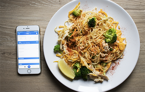 A plate of noodles and broccoli next to a cell phone