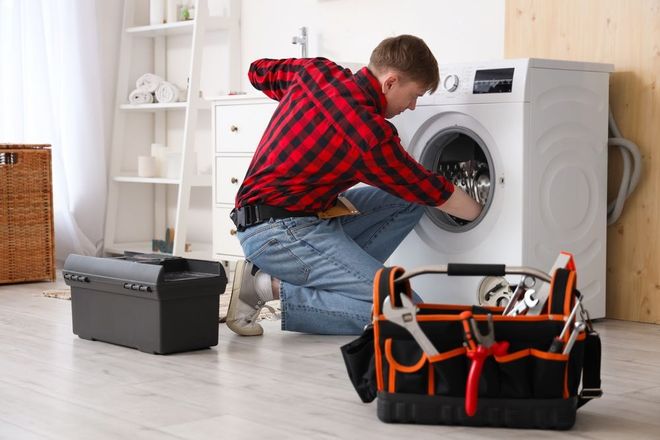 Man in red plaid shirt repairs a washing machine, tools in front. Light-filled room.