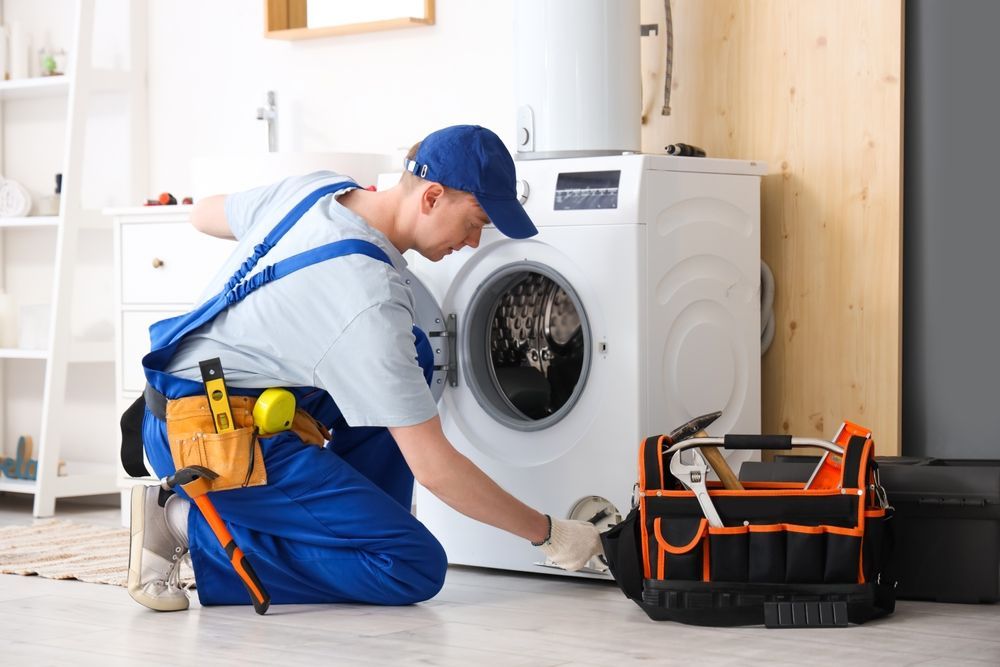 A repairman in blue overalls examines a washing machine, tools at his side, in a laundry room.