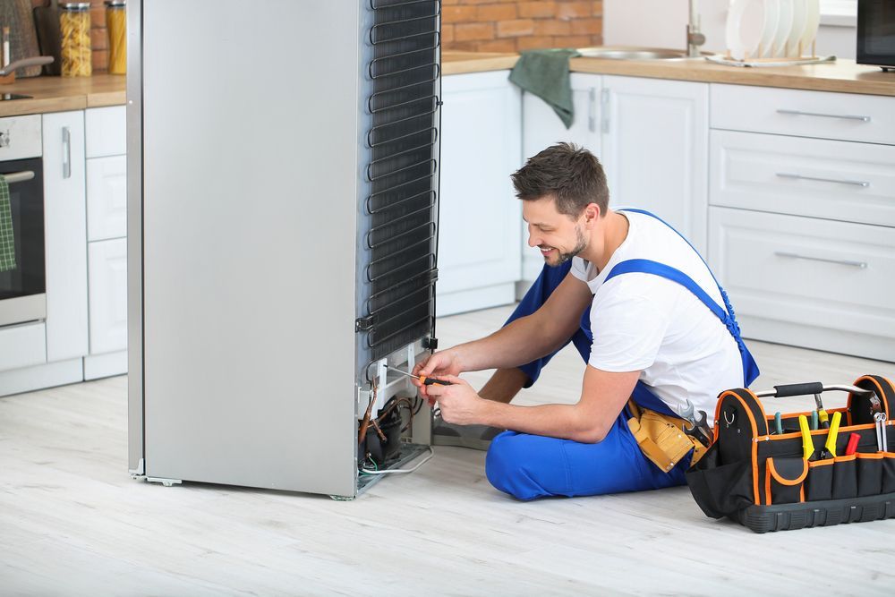Refrigerator repairman working on the back of the appliance in a kitchen, smiling. Toolbox nearby.