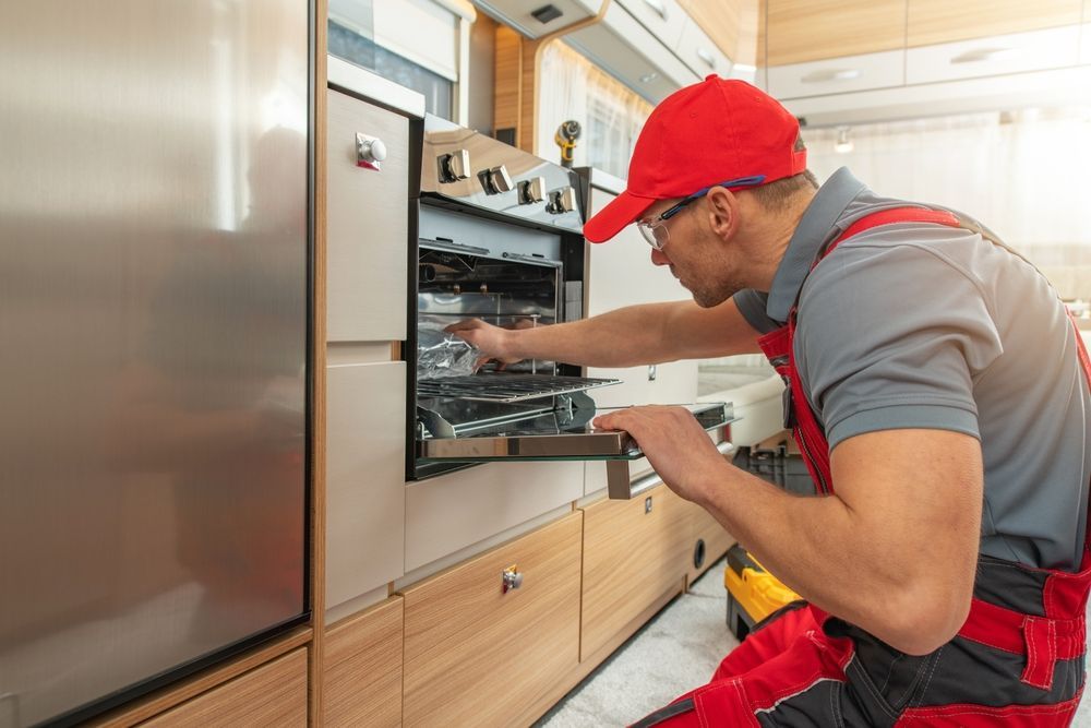 A person in red overalls and hat repairs an oven inside a camper.