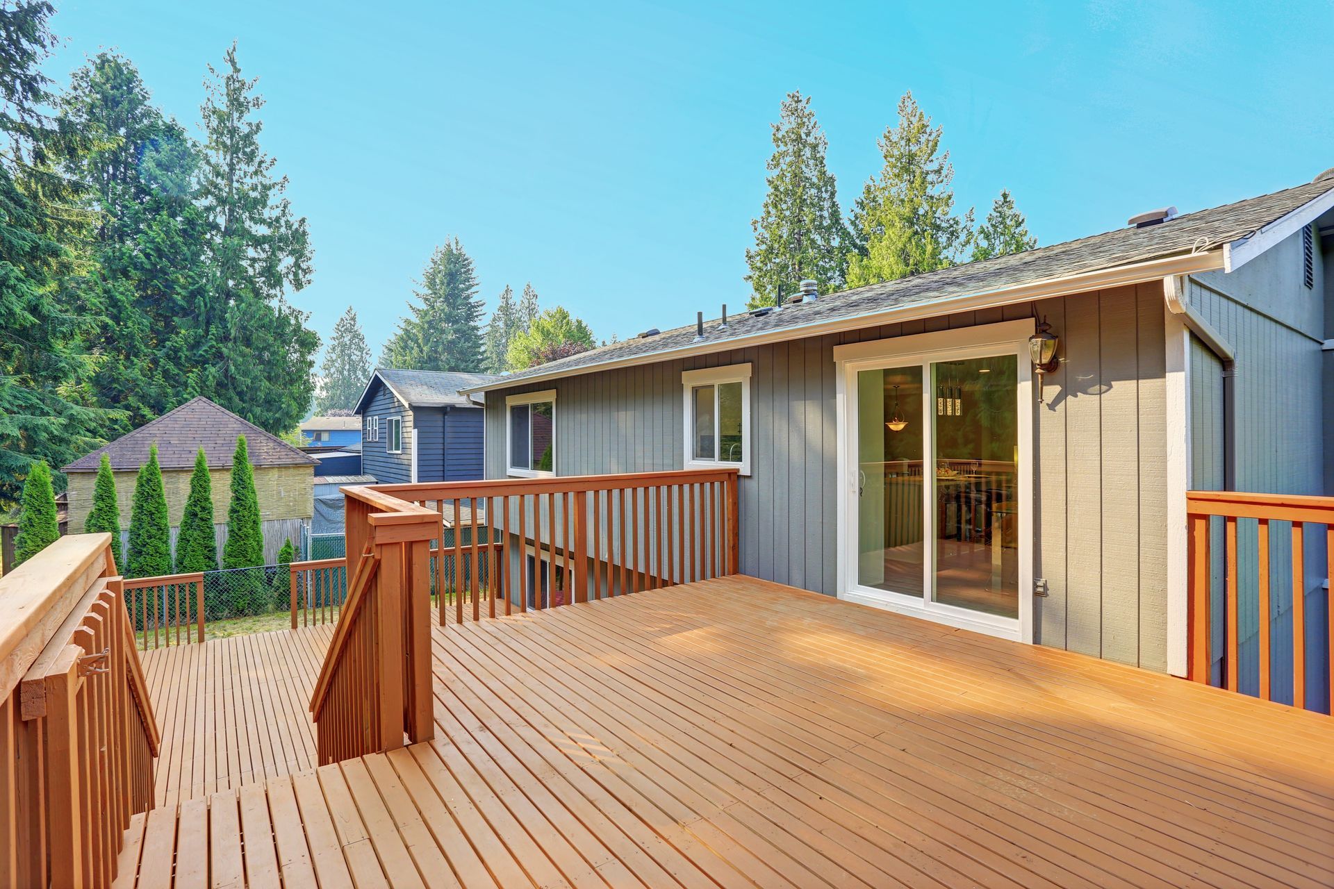 Wooden deck connected to a gray house with sliding glass doors and surrounded by trees under a blue sky.