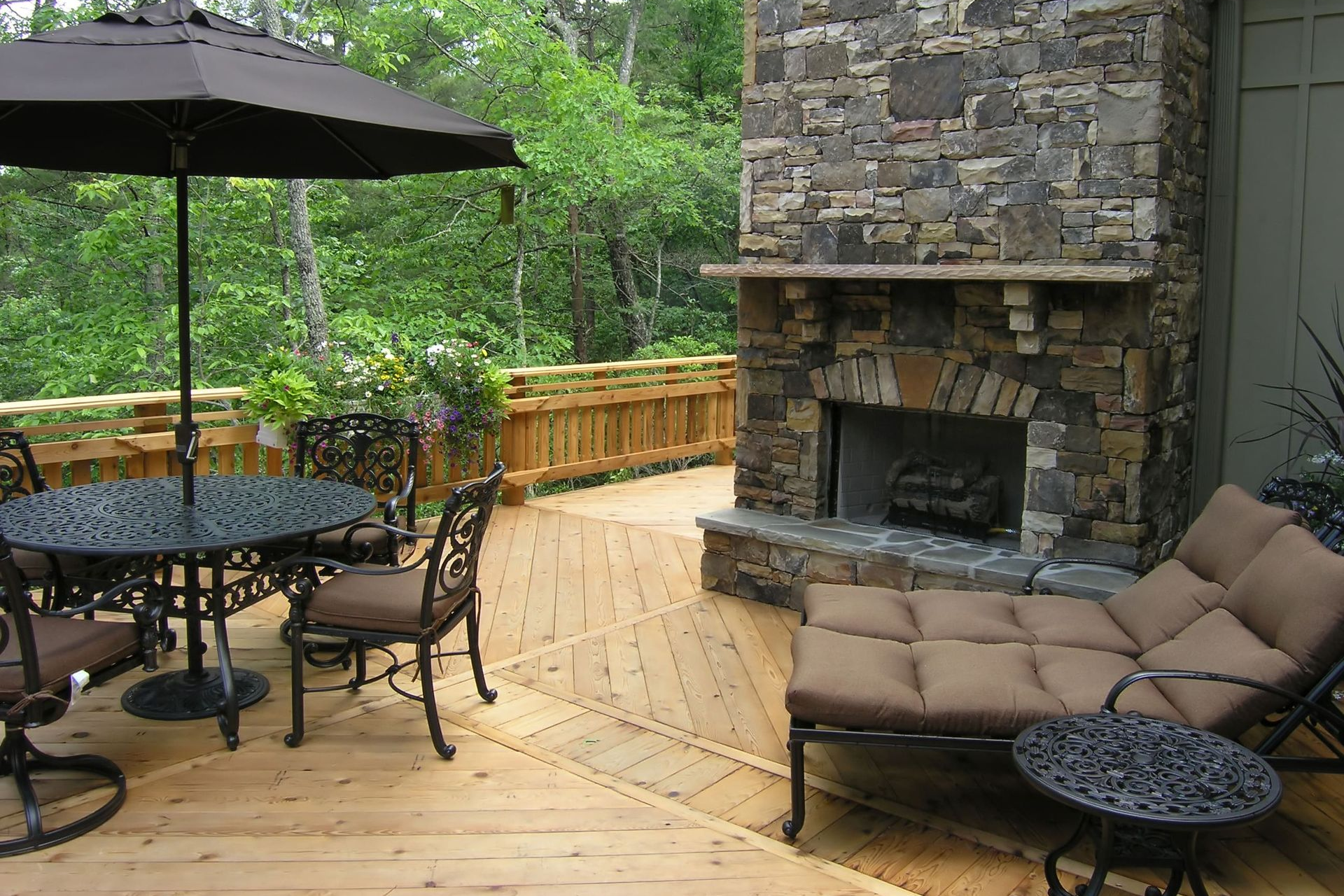 Outdoor deck with furniture, stone fireplace, and umbrella, surrounded by trees.