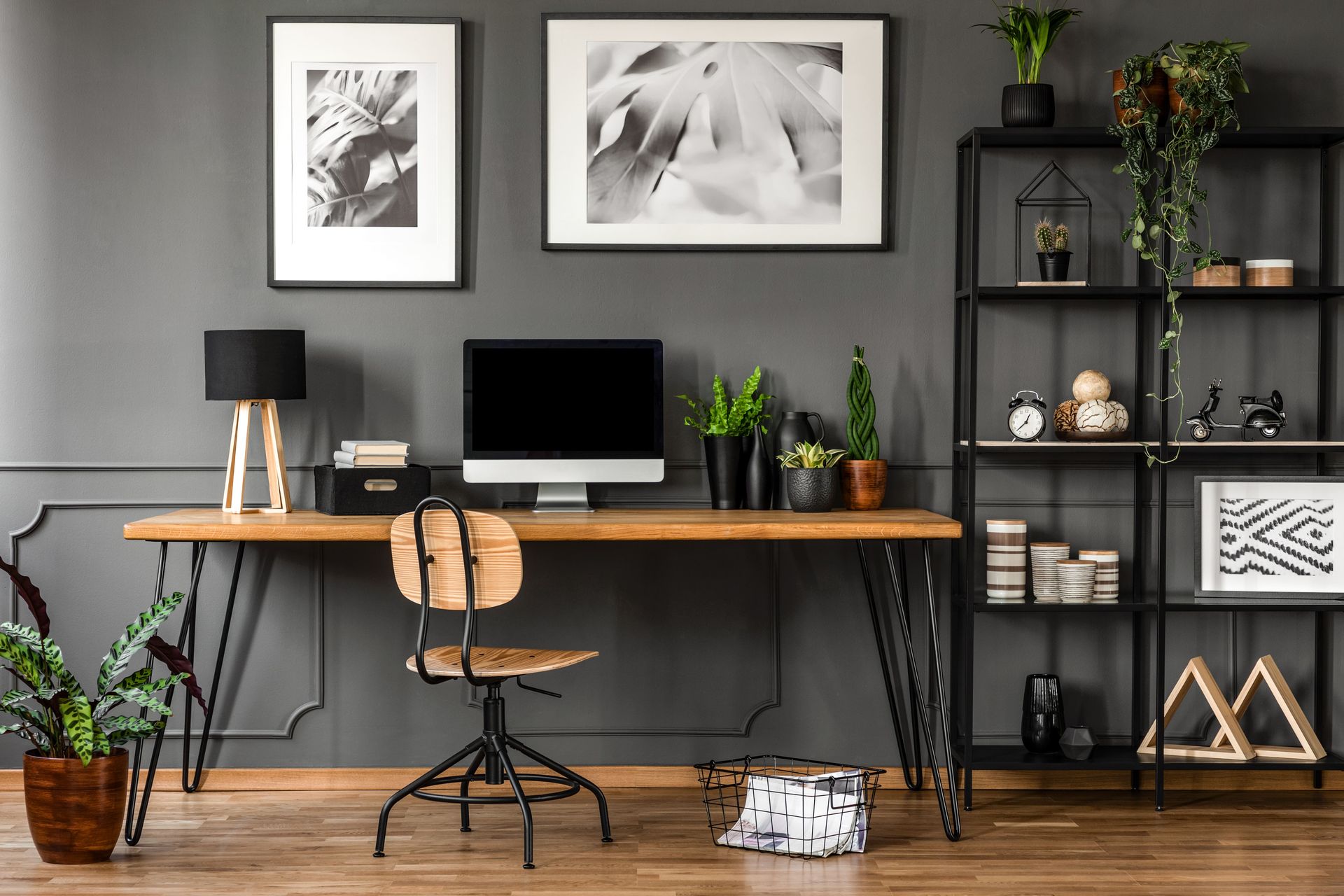 Home office with a wooden desk, black chair, and metal shelving. Grey wall, black and white artwork, plants.