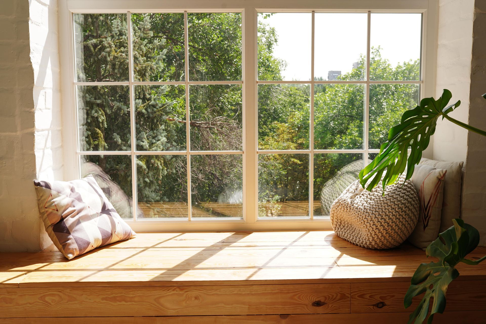 Window seat with sunlight, overlooking trees. Cushions and a plant rest on the wooden ledge.