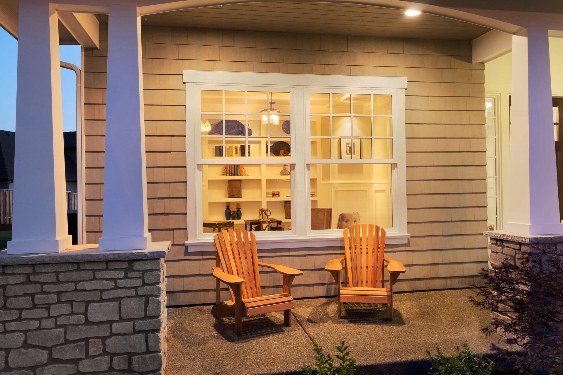 Two wooden Adirondack chairs sit on a porch in front of a brightly lit window at dusk.