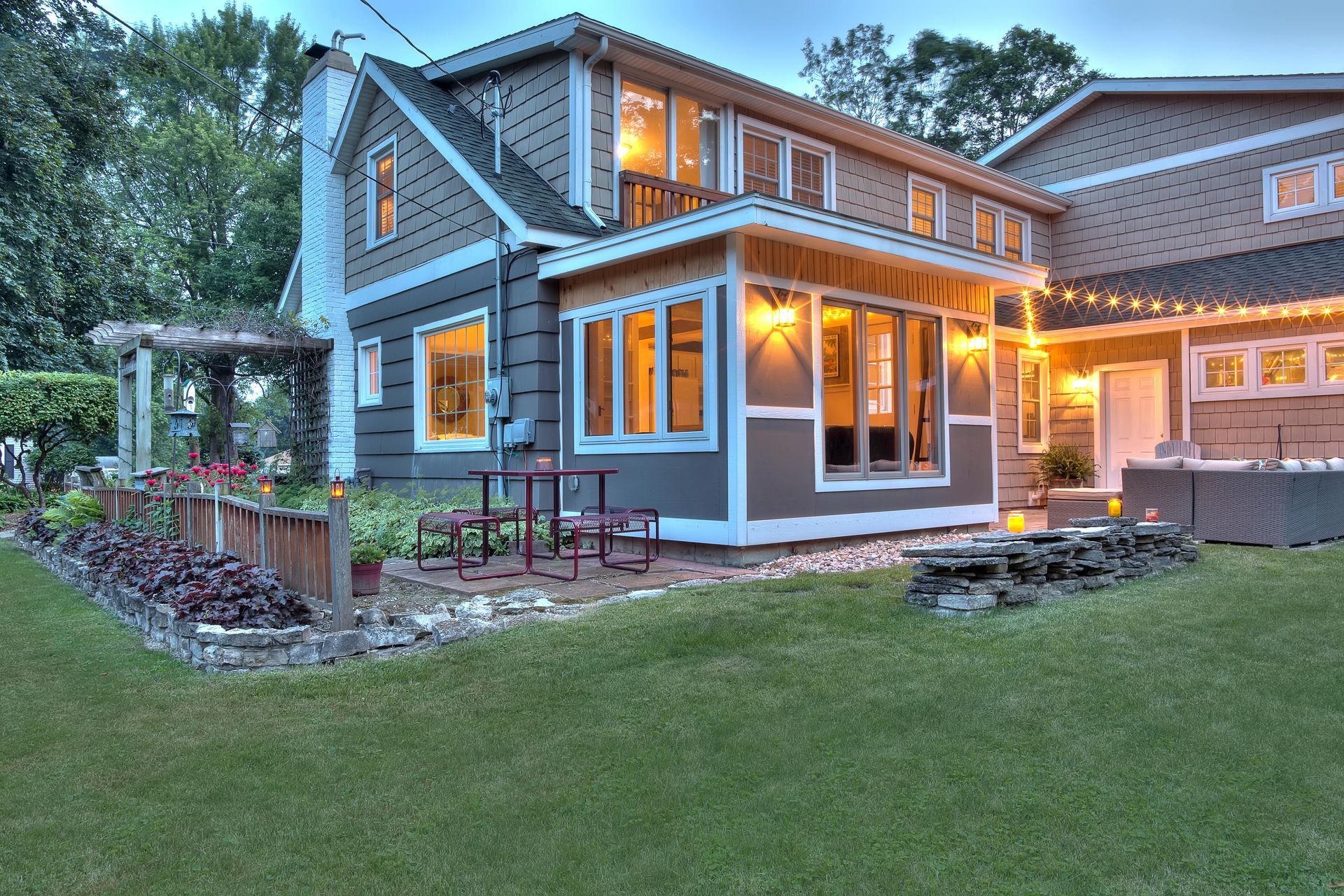 Two-story house with dark grey siding, lit windows, patio with table and chairs, and a hot tub.