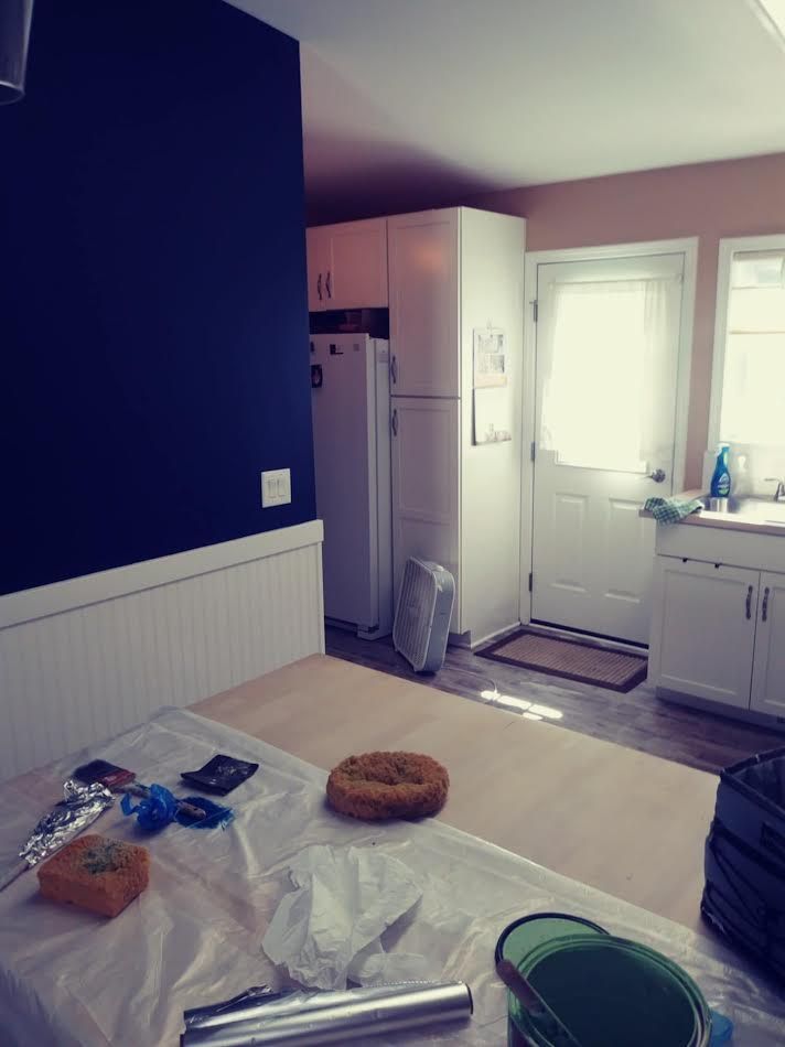 Kitchen interior with a blue wall, white cabinets, a table covered in items, and a white door.
