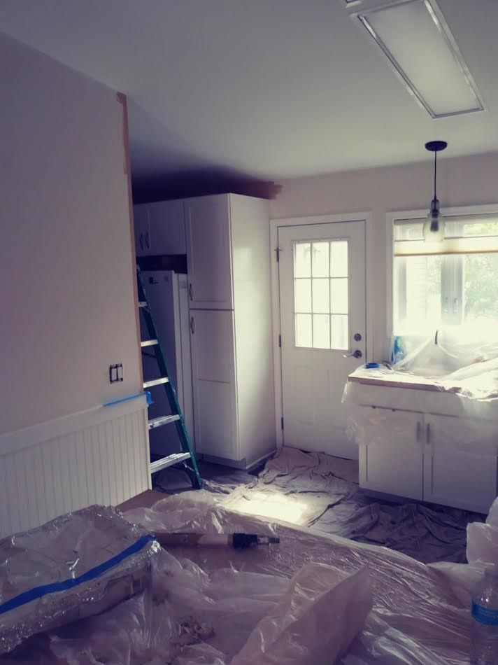 Kitchen under renovation: white cabinets, door, and fridge; ladder; plastic sheeting covers floor.