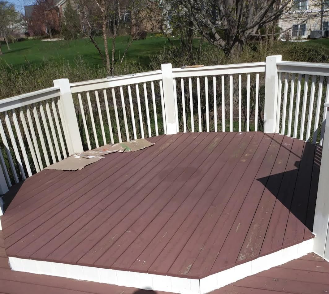Brown wooden deck with white railing, surrounded by grass and trees on a sunny day.