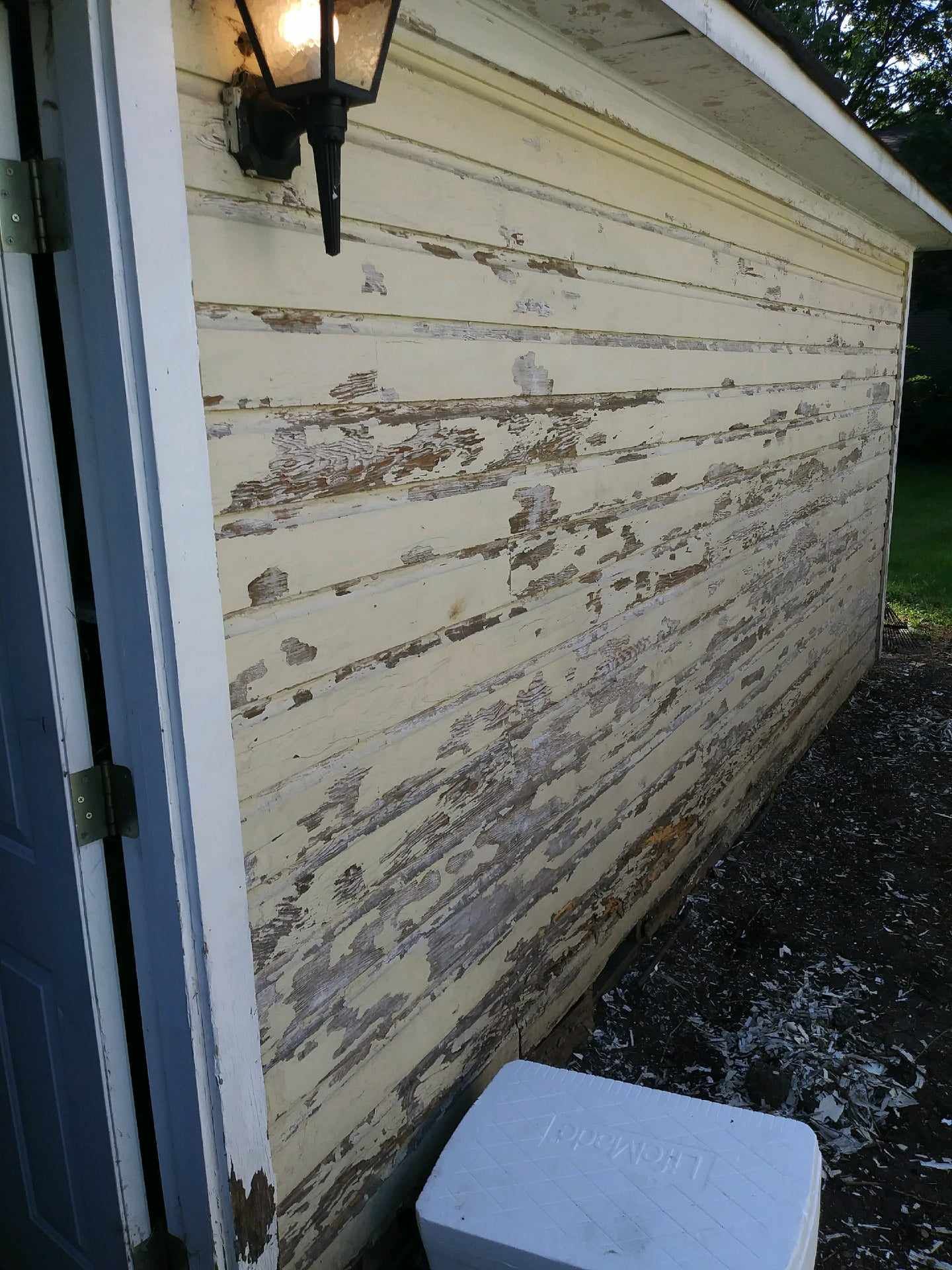 Weathered yellow siding on a building, with peeling paint. A light is mounted above. A white cooler sits below.